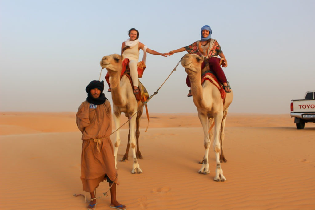 Two women jooin hands while riding atop separate camels