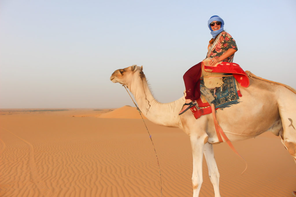 A woman rides on a camel in colorful garbs and sunglasss