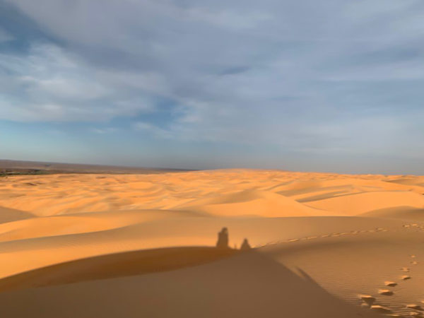 Shadow figures appear from the top of a large dune cast by a setting sun